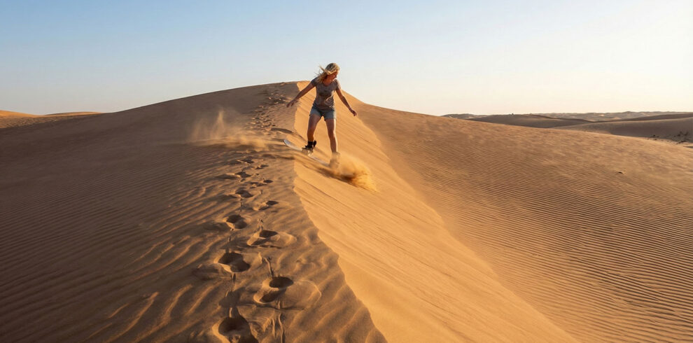 Un viaggiatore che fa sandboarding scivolando lungo le alte dune di sabbia dorata del deserto di Wahiba Sands, Oman
