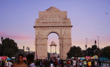 A bustling scene at India Gate with a vibrant crowd under a colorful sunset sky.