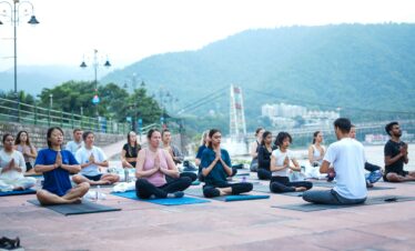 A diverse group practicing yoga by the river in Rishikesh, India, surrounded by scenic mountains.