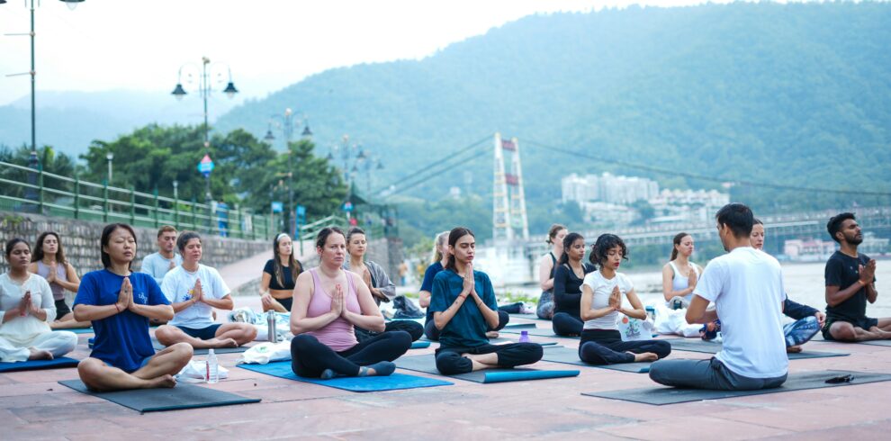 A diverse group practicing yoga by the river in Rishikesh, India, surrounded by scenic mountains.